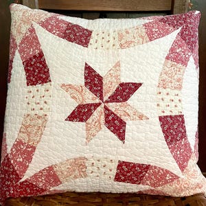 May include: Square quilted pillow with a central star design. The star is composed of red and white patterned fabric, surrounded by a white background and red patterned borders. The pillow is placed on a wooden chair.