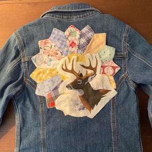 May include: Blue denim jacket with a colorful patchwork design on the back. The patchwork features a deer illustration and various fabric patterns in shades of blue, yellow, red, and white. The jacket has a classic collar and button-up front.