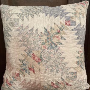 May include: Square quilted pillow with a pineapple design. The pillow features a patchwork pattern with floral and geometric elements in shades of white, blue, and pink. The pillow is placed on a wooden chair.