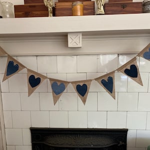 May include: A rustic banner with triangular burlap flags strung on twine. Each flag features a heart, some in denim and others in a darker blue fabric. The banner is displayed above a fireplace.