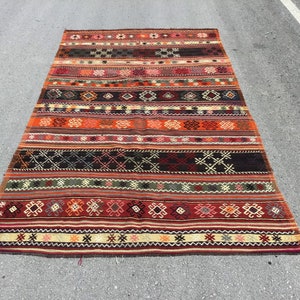 May include: A traditional Turkish kilim rug with a colorful geometric pattern in shades of red, orange, green, and black. The rug has a long, rectangular shape and is woven with intricate details.