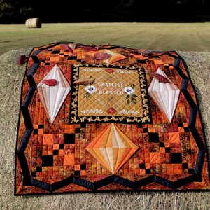May include: A quilted wall hanging with a patchwork design in shades of orange, brown, and black. The center features a rectangular panel with the text "Grateful Blessed" in a gold and white font. The quilt is displayed on a bale of hay.