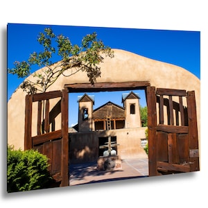 May include: A photograph of a historic adobe church with wooden doors and a clear blue sky. The church has a tan exterior with a wooden door frame. A tree grows over the top of the door frame. The doors are open, revealing the church's interior.