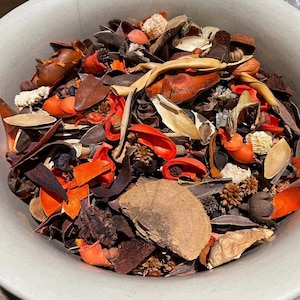 May include: A white ceramic bowl filled with a colorful assortment of dried potpourri. The potpourri includes various shapes and textures, with shades of orange, brown, beige, and red. The bowl is round and has a simple, clean design.