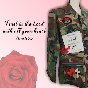 May include: A camouflage jacket with red and white embroidered hearts, roses, and a cross. The jacket has a patch with the text "Trust in the Lord with all your heart. Proverbs 3:5".