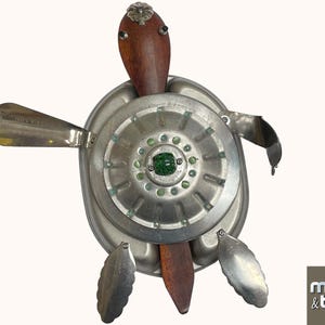 May include: A handcrafted metal turtle sculpture with a wooden head and legs. The turtle's shell is made from repurposed metal parts, with a central circular element and green accents. The sculpture is signed "mutts & bolts".