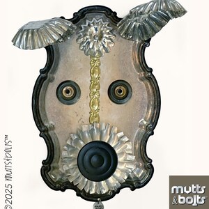 May include: A unique, handcrafted metal art piece resembling a dog's face. It features a silver-colored tray as the base, with decorative elements like a gold-colored stem, silver-colored flower shapes, and a black speaker. The piece includes the text "©2025 Mutts & Bolts™".