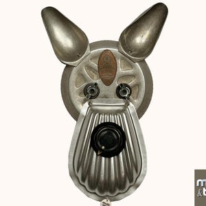 May include: A silver metal sculpture of a dog's head, made from repurposed kitchen utensils. The dog's ears are made from two silver metal spoons, the nose is a silver metal pie plate, and the eyes are two silver metal knobs. The sculpture is titled "Mutt & Bolts" and is signed with the year 2024.