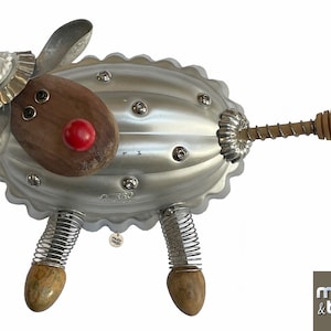 May include: A whimsical metal sheep sculpture made from repurposed materials. The body is a silver, scalloped-edged form with rivets. It features wooden elements for the head and legs, a red nose, and spring legs. The tail has a wooden handle with a decorative end.