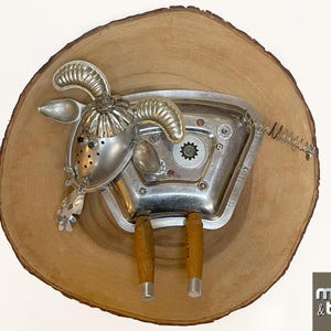 May include: A unique metal sculpture of a ram, crafted from repurposed silverware and other metal objects. The ram is mounted on a round wooden plaque. The sculpture features a spring for a tail and knife handles for legs.