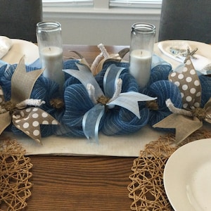May include: A blue mesh table runner with decorative bows, candles, and placemats. The bows have tan and white polka dot accents. White candles are in glass holders. White plates and napkins are set on the table.
