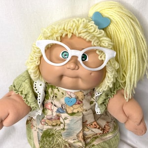 May include: A vintage doll with yellow yarn hair styled in a ponytail and curls, wearing white cat-eye glasses. The doll has green eyes and is dressed in a green and patterned dress with a blue heart-shaped button.