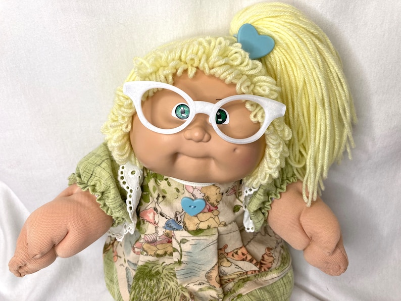 May include: A vintage doll with yellow yarn hair styled in a ponytail and curls, wearing white cat-eye glasses. The doll has green eyes and is dressed in a green and patterned dress with a blue heart-shaped button.