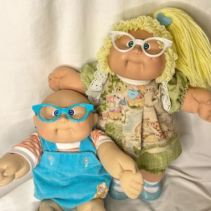May include: Two vintage Cabbage Patch Kids dolls. One doll wears blue corduroy dungarees and blue glasses. The other doll wears a green dress with a Winnie the Pooh print and white glasses. Both dolls have soft, vinyl faces.