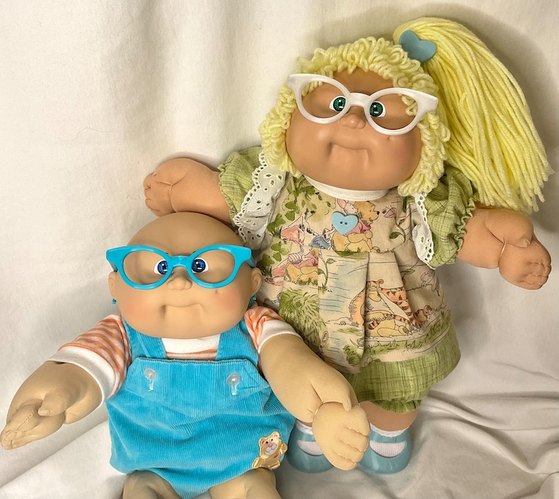 May include: Two vintage Cabbage Patch Kids dolls. One doll wears blue corduroy dungarees and blue glasses. The other doll wears a green dress with a Winnie the Pooh print and white glasses. Both dolls have soft, vinyl faces.