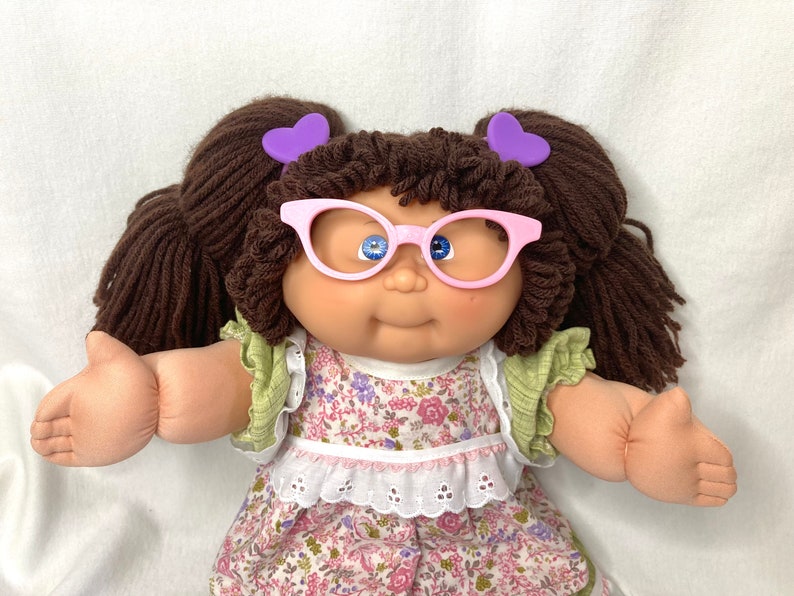 May include: A vintage doll with brown yarn pigtails, decorated with purple heart-shaped hair clips. The doll wears pink-rimmed glasses and a floral dress with green sleeves. The doll's skin is a light peach colour.