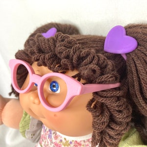 May include: A doll with brown yarn hair, blue eyes, and pink glasses. The doll has two purple heart-shaped hair clips. The doll is wearing a floral print dress with a green top.