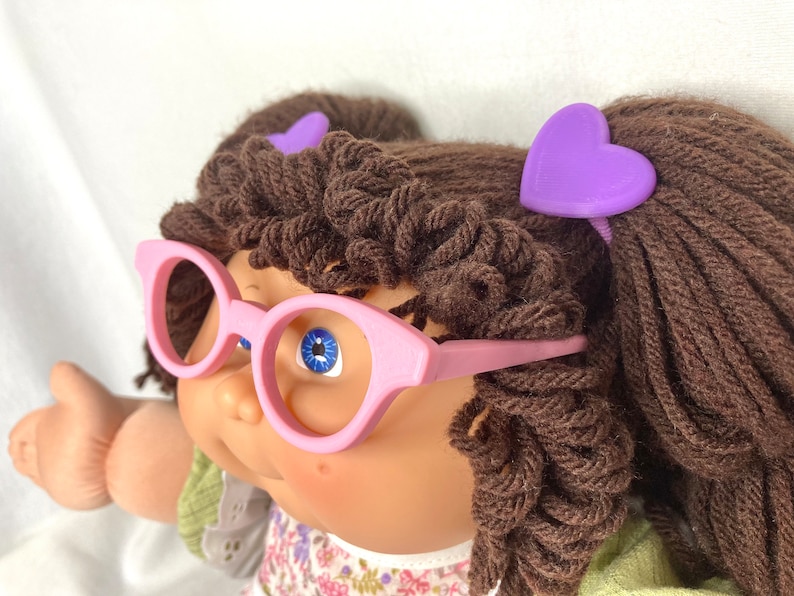 May include: A doll with brown yarn hair, blue eyes, and pink glasses. The doll has two purple heart-shaped hair clips. The doll is wearing a floral print dress with a green top.
