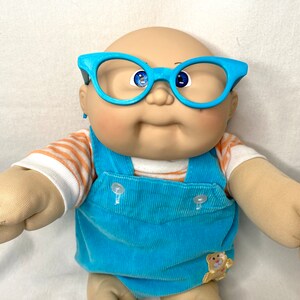 May include: A Cabbage Patch Kid doll wearing bright blue glasses, a blue corduroy overall, and a striped orange and white shirt. The doll has blue eyes and a small teddy bear patch on the overall.