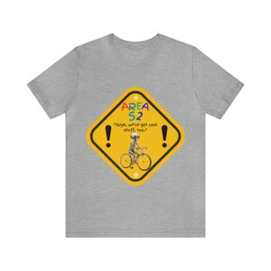 May include: A gray t-shirt with a yellow diamond-shaped sign that says "AREA 52" in rainbow colors. The sign also says "Guys, we've got cool stuff, too." and has an image of a gray alien riding a bicycle.