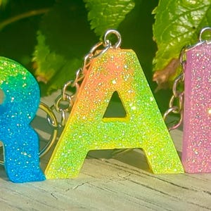May include: Three glittery resin keychains spelling out the word "RAD". The letters are in rainbow colors, with the "R" transitioning from orange to blue, the "A" from yellow to green, and the "D" in pink. Each letter has a silver chain.