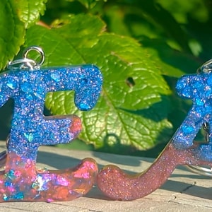 May include: Five resin letter charms with glitter embedded in the resin. The letters spell out the word "NEAJ". The letters are in shades of blue, pink, and green.