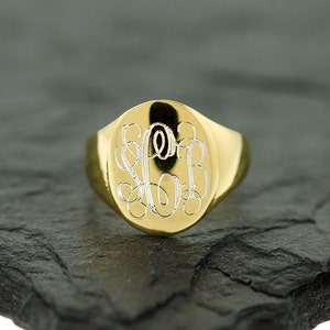 May include: A gold signet ring with an oval-shaped top featuring a monogram engraved in white.