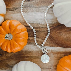 May include: A white pearl necklace with a silver toggle clasp and a round silver charm with the initials "JM" engraved on it. The necklace is displayed on a wooden surface with several white and orange pumpkins.