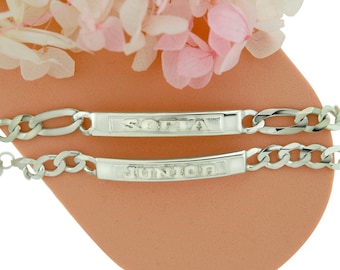 May include: Two silver-toned bracelets with a chain link design. Each bracelet has a rectangular plate with the names "SOFIA" and "JUNIOR" engraved. The bracelets are displayed on a peach-colored surface, with pink and white flowers in the background.