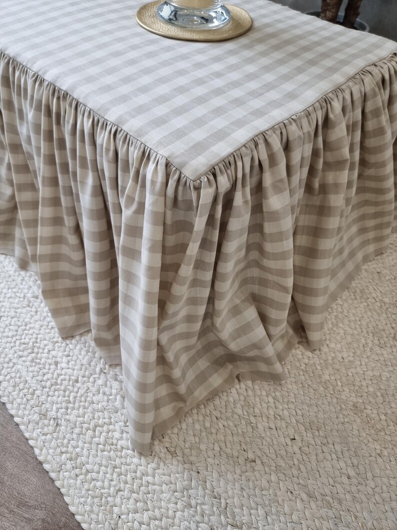 May include: A beige and white checkered table with a ruffled skirt. The table is covered with a fabric featuring a classic gingham pattern. The skirt adds a decorative element to the table's design.
