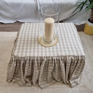 May include: A square ottoman with a beige and white checkered fabric cover and a ruffled skirt. A clear glass hurricane lantern with a beige candle sits on top of the ottoman. The ottoman is on a woven rug.