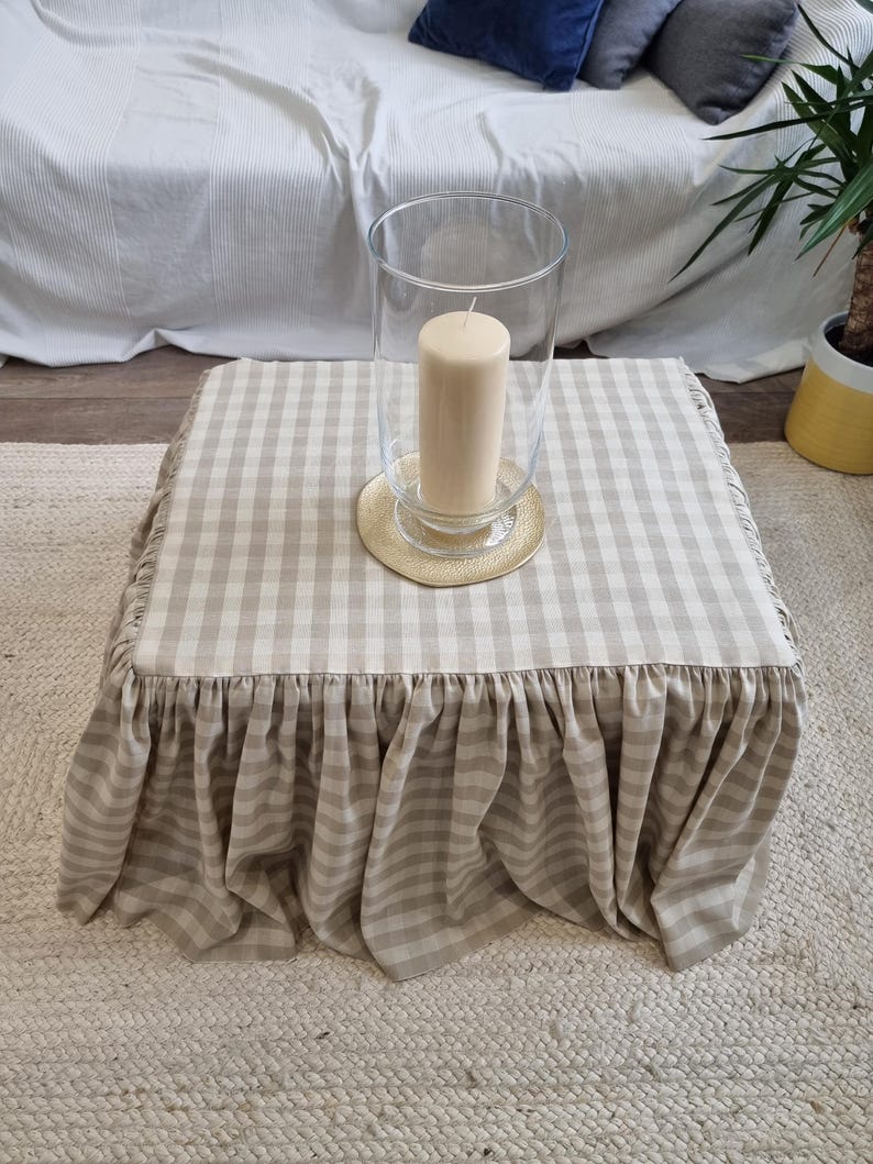 May include: A square ottoman with a beige and white checkered fabric cover and a ruffled skirt. A clear glass hurricane lantern with a beige candle sits on top of the ottoman. The ottoman is on a woven rug.