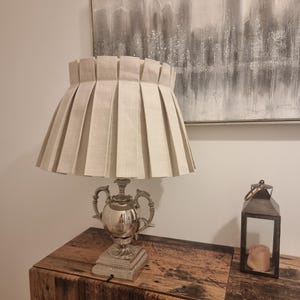 May include: A table lamp featuring a metallic silver base with ornate curved handles and a square pedestal. The lamp is topped with a wide, beige fabric lampshade characterised by deep, vertical box pleats that create a structured, uniform appearance.