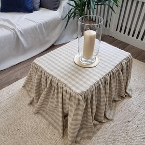 May include: A square coffee table with a beige and white checkered top and a ruffled skirt. A large glass candle holder with a beige candle sits on the table. The table is on a cream-colored rug.