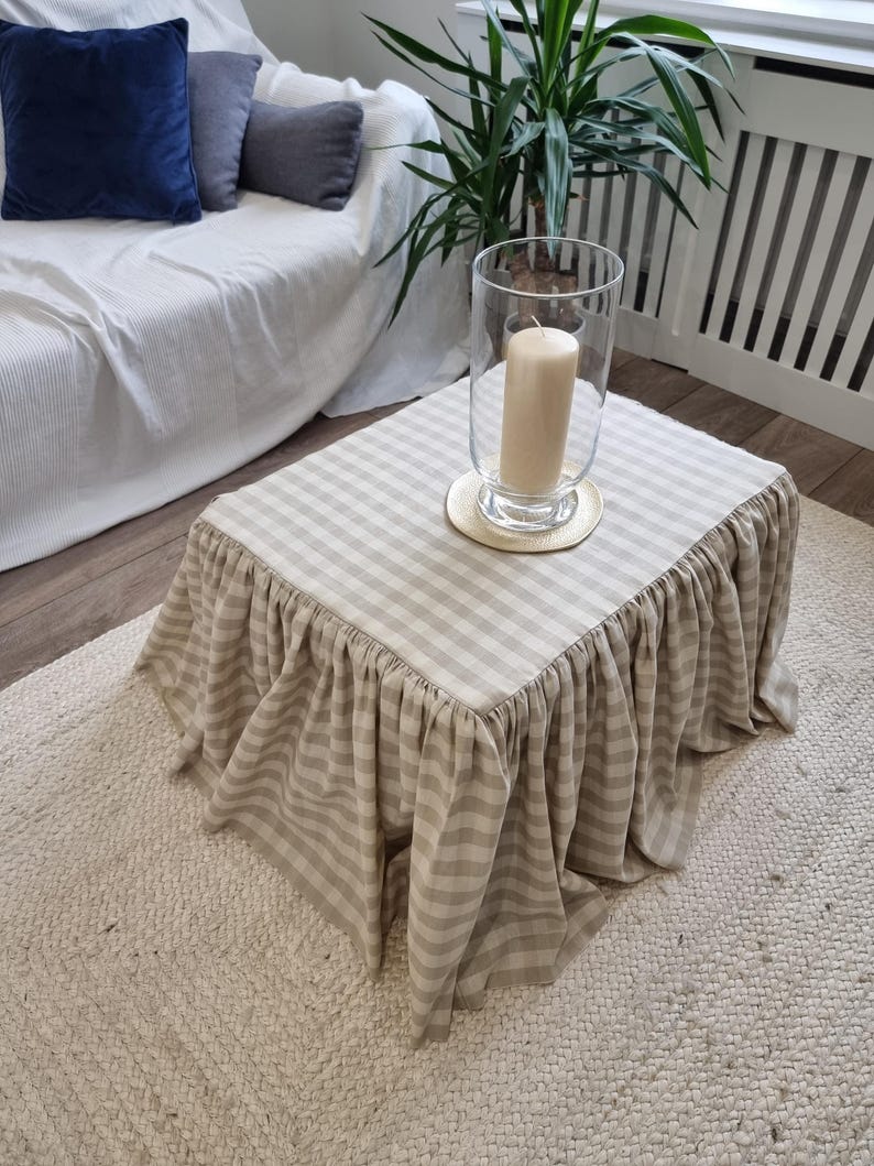May include: A square coffee table with a beige and white checkered top and a ruffled skirt. A large glass candle holder with a beige candle sits on the table. The table is on a cream-colored rug.