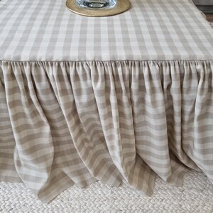 May include: A beige and white checkered table cover with a ruffled skirt. The table cover is draped over a square table. A glass candle holder with a gold base sits on top of the table.