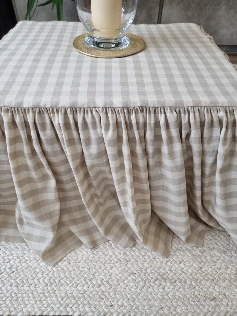 May include: A beige and white checkered table cover with a ruffled skirt. The table cover is draped over a square table. A glass candle holder with a gold base sits on top of the table.