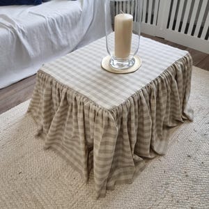 May include: A small square coffee table with a beige and white striped skirt. A large clear glass hurricane lamp with a beige candle sits on top of the table. The table is on a woven rug.