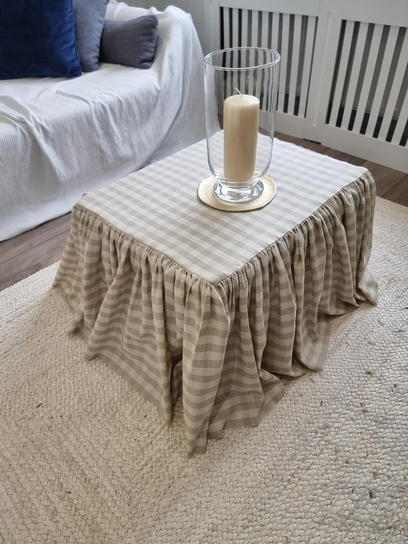 May include: A small square coffee table with a beige and white striped skirt. A large clear glass hurricane lamp with a beige candle sits on top of the table. The table is on a woven rug.