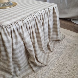 May include: A beige and white striped ottoman with a ruffled skirt. The fabric has a classic gingham pattern. The ottoman sits on a woven jute rug. A glass vase and gold plate are visible on the ottoman's surface.