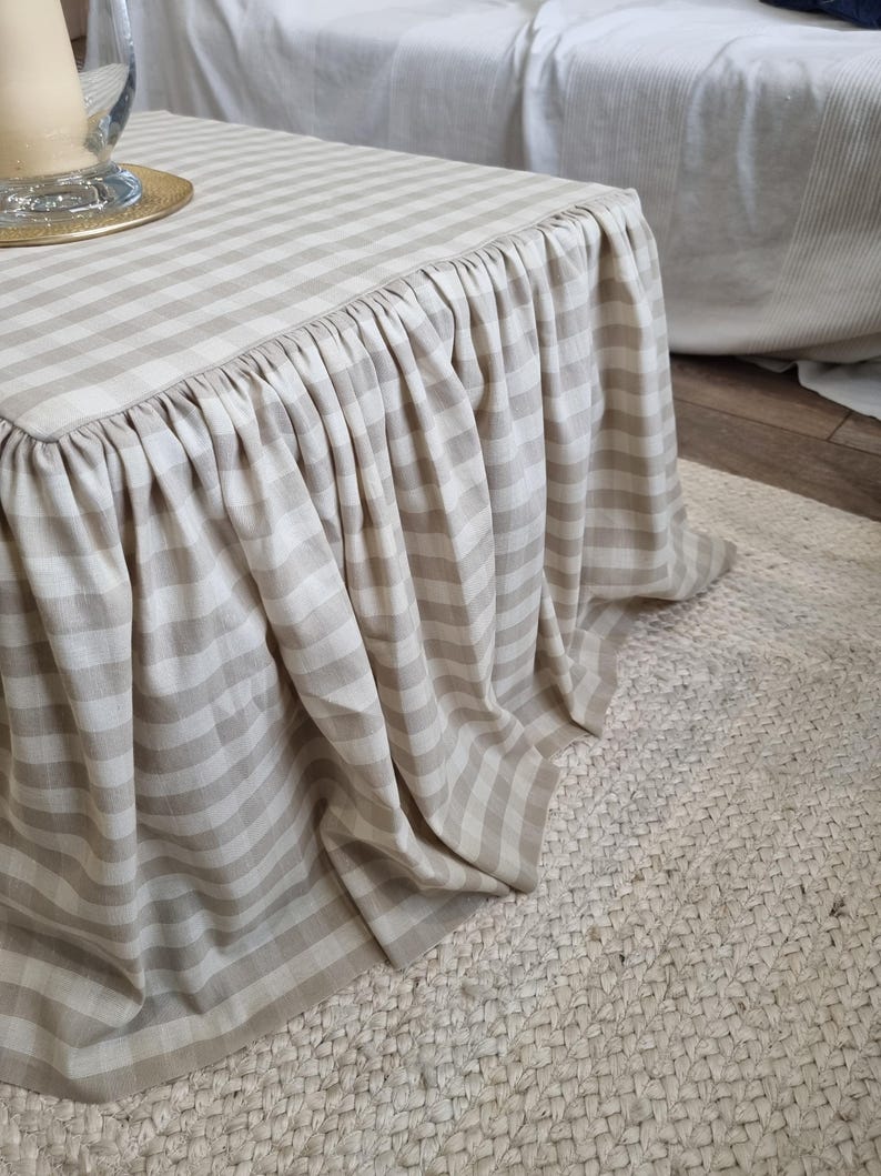 May include: A beige and white striped ottoman with a ruffled skirt. The fabric has a classic gingham pattern. The ottoman sits on a woven jute rug. A glass vase and gold plate are visible on the ottoman's surface.