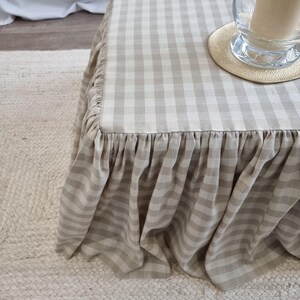 May include: A square ottoman with a beige and white gingham pattern fabric skirt. The skirt has a ruffled edge. A clear glass vase with a candle and a small round coaster sit on top of the ottoman.