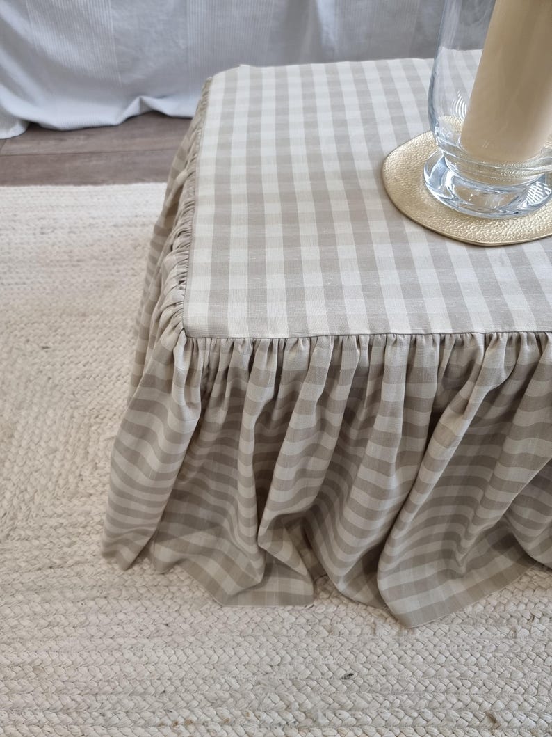 May include: A square ottoman with a beige and white gingham pattern fabric skirt. The skirt has a ruffled edge. A clear glass vase with a candle and a small round coaster sit on top of the ottoman.