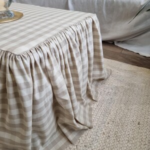 May include: A beige and white striped table cover with a ruffled skirt. The cover is draped over a square table, with the stripes running horizontally. The table sits on a woven jute rug, and a white sofa is in the background.