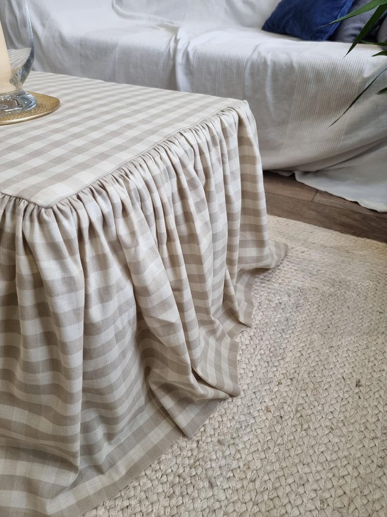 May include: A beige and white striped table cover with a ruffled skirt. The cover is draped over a square table, with the stripes running horizontally. The table sits on a woven jute rug, and a white sofa is in the background.