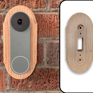 May include: A wooden door frame with a rounded top and a cutout for a doorbell. The doorbell is a gray, rectangular device with a round camera lens and a white circle around the lens. The frame is made of light brown wood and has a natural wood finish.