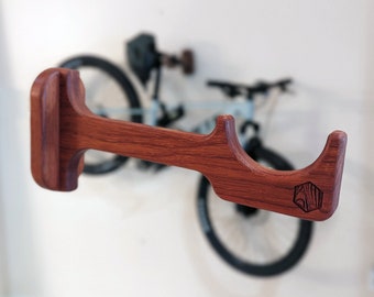 May include: A wooden bike rack with a reddish-brown finish. The rack has a curved design and a logo on the side. The rack is mounted on a white wall.