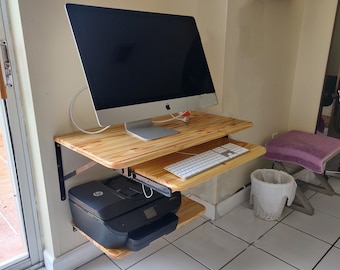 May include: A wooden desk with a black computer monitor, a white keyboard, and a black printer. The desk is mounted to the wall with black brackets.
