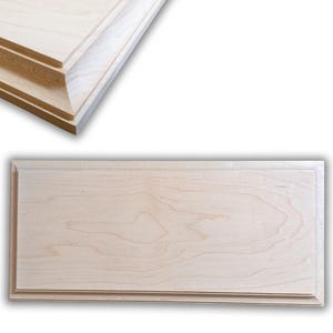 May include: A light-colored, rectangular wooden plaque with a raised border. The wood grain is visible, and the plaque is unfinished. A corner of a similar plaque is visible in the upper left.