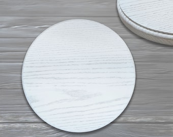 Puede incluir: Dos posavasos redondos de madera blanca con un patrón de veta de madera. Los posavasos están sobre una superficie de madera gris.